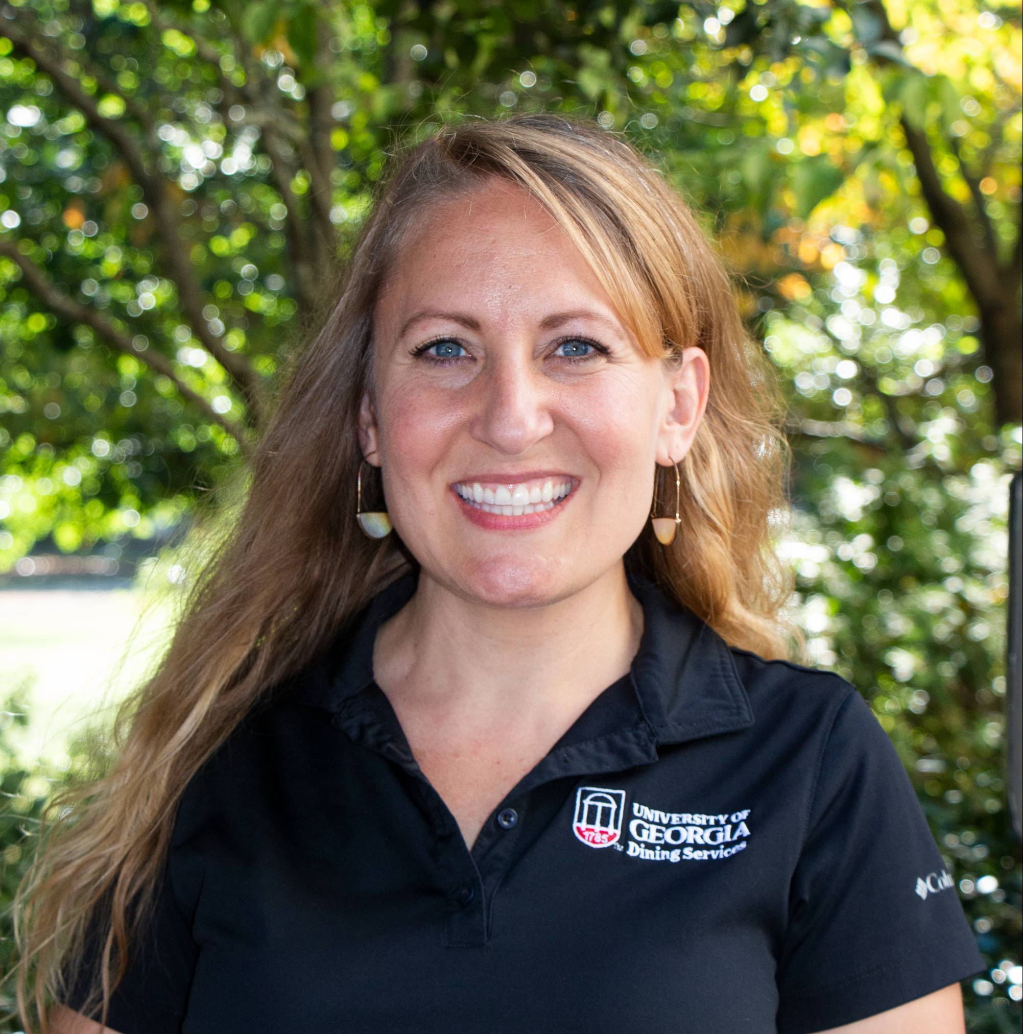 A woman with long blonde hair wearing gold earings and a black shirt with the words "University of Georgia, Dining Services" smiles at the camera.