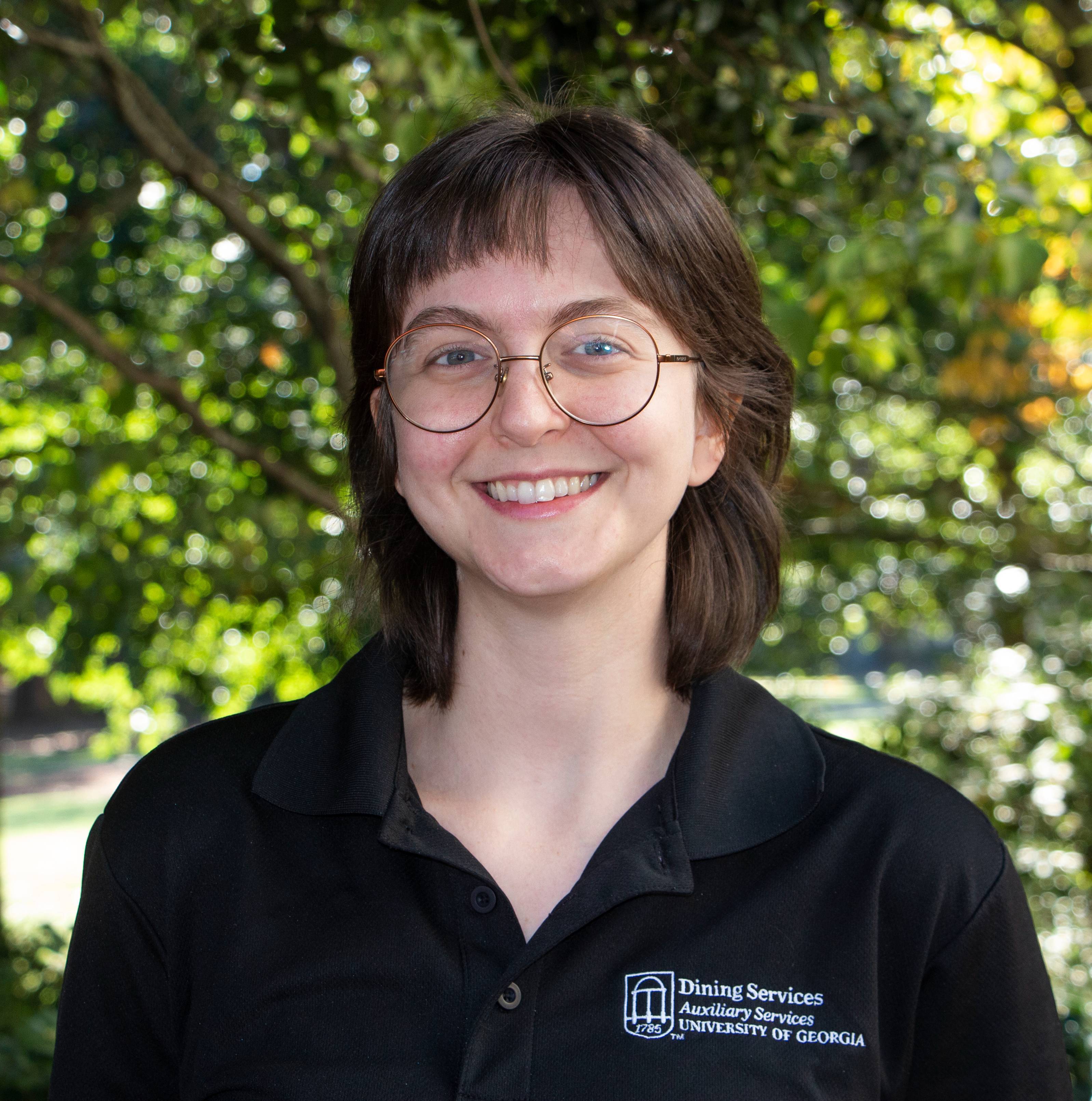 A woman with shoulder-length brown hair wearing round frame glasses and a black shirt with the words "Dining Services, Auxiliary Services, University of Georgia" smiles at the camera.