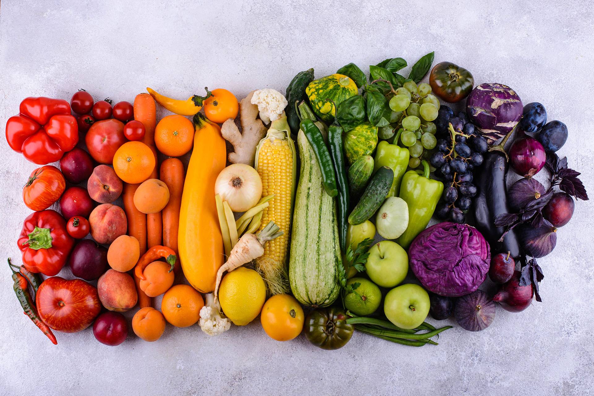 an assortment of fruits and vegetables, arranged in ranbow order on a grey background/