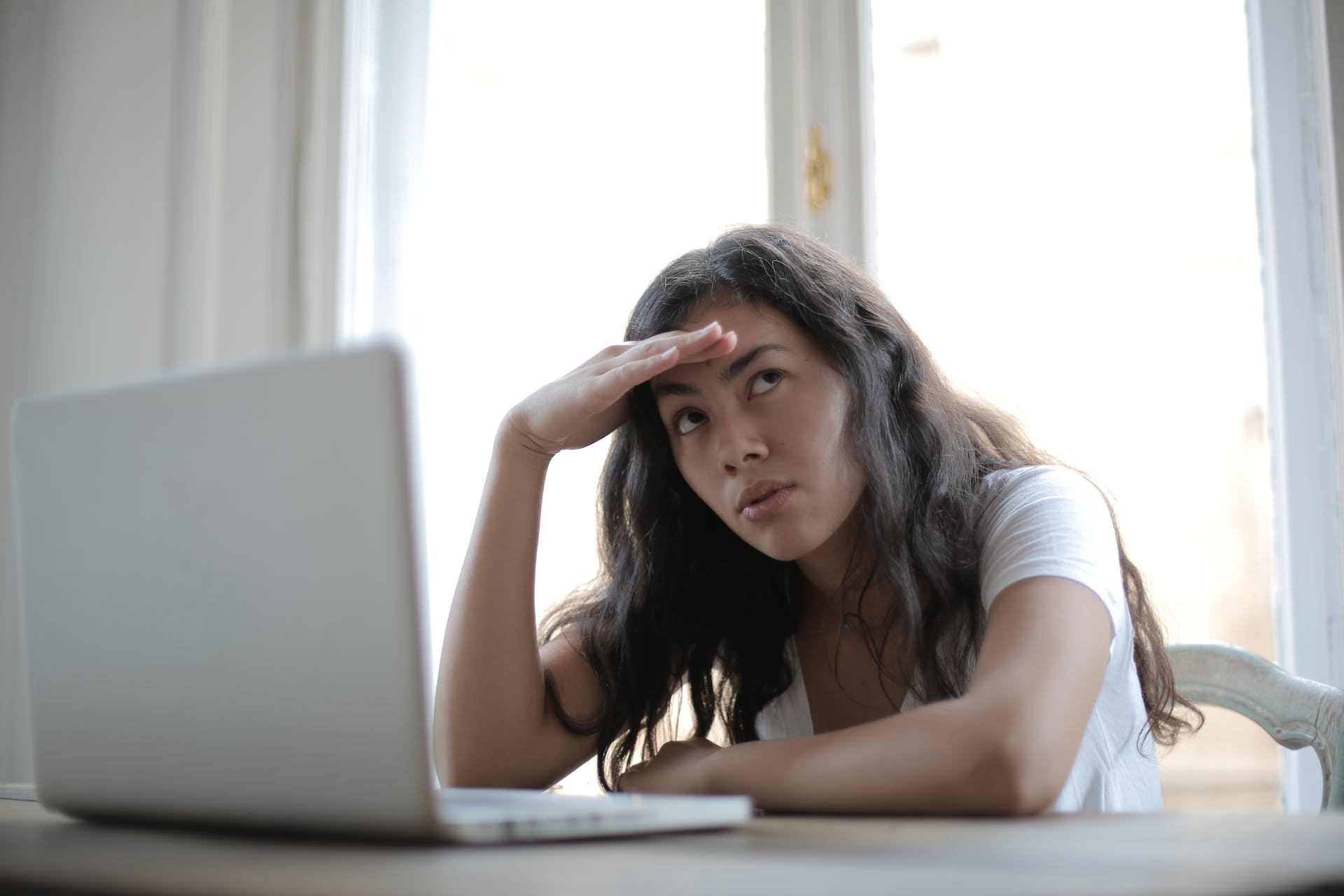A photograph of a woman sitting in front of a laptop and holding her head while looking to the side as if trying to remember something.