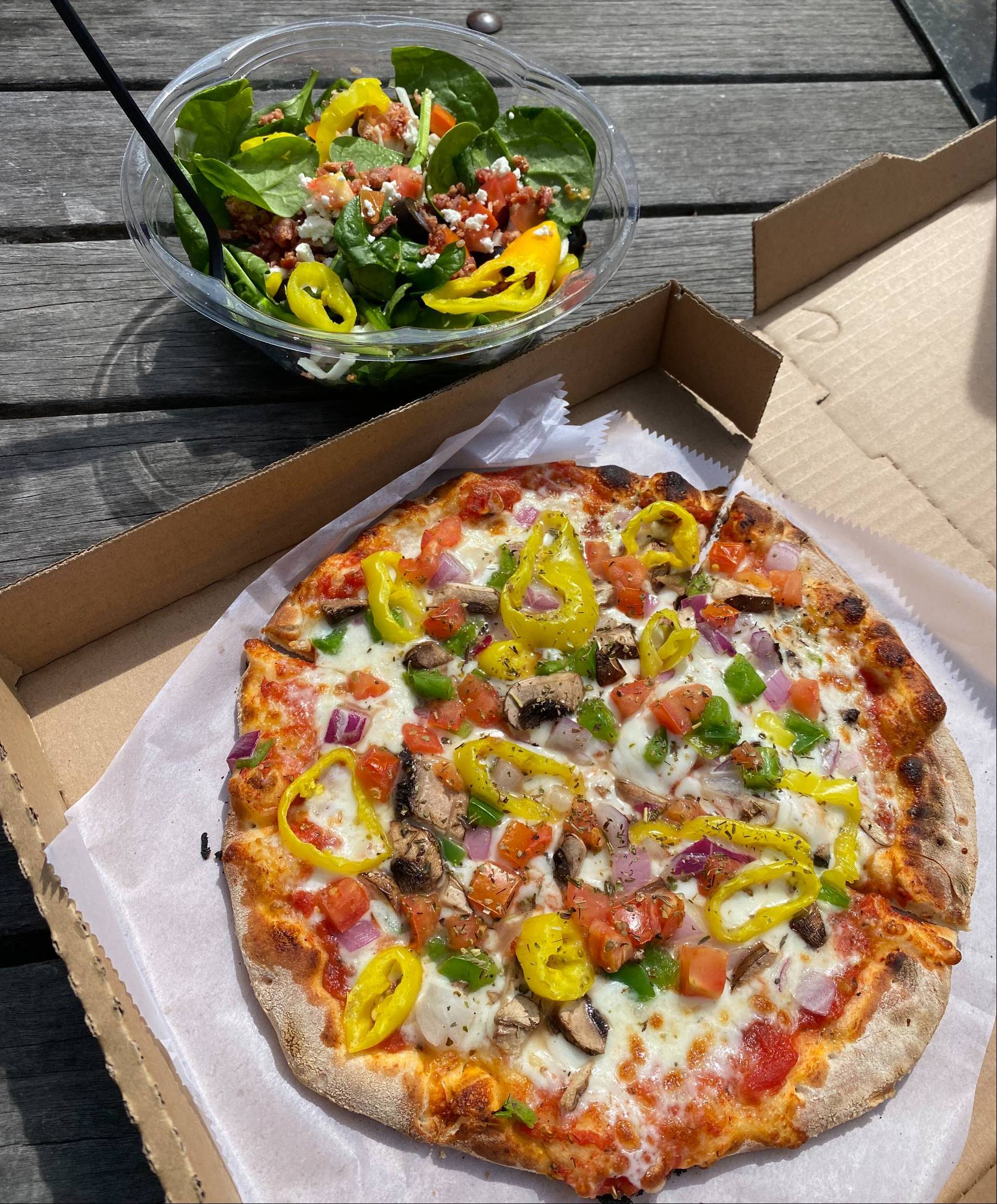 A photograph of a pizza in a box sitting on a picnic table with a side salad sitting in a bowl beside it.