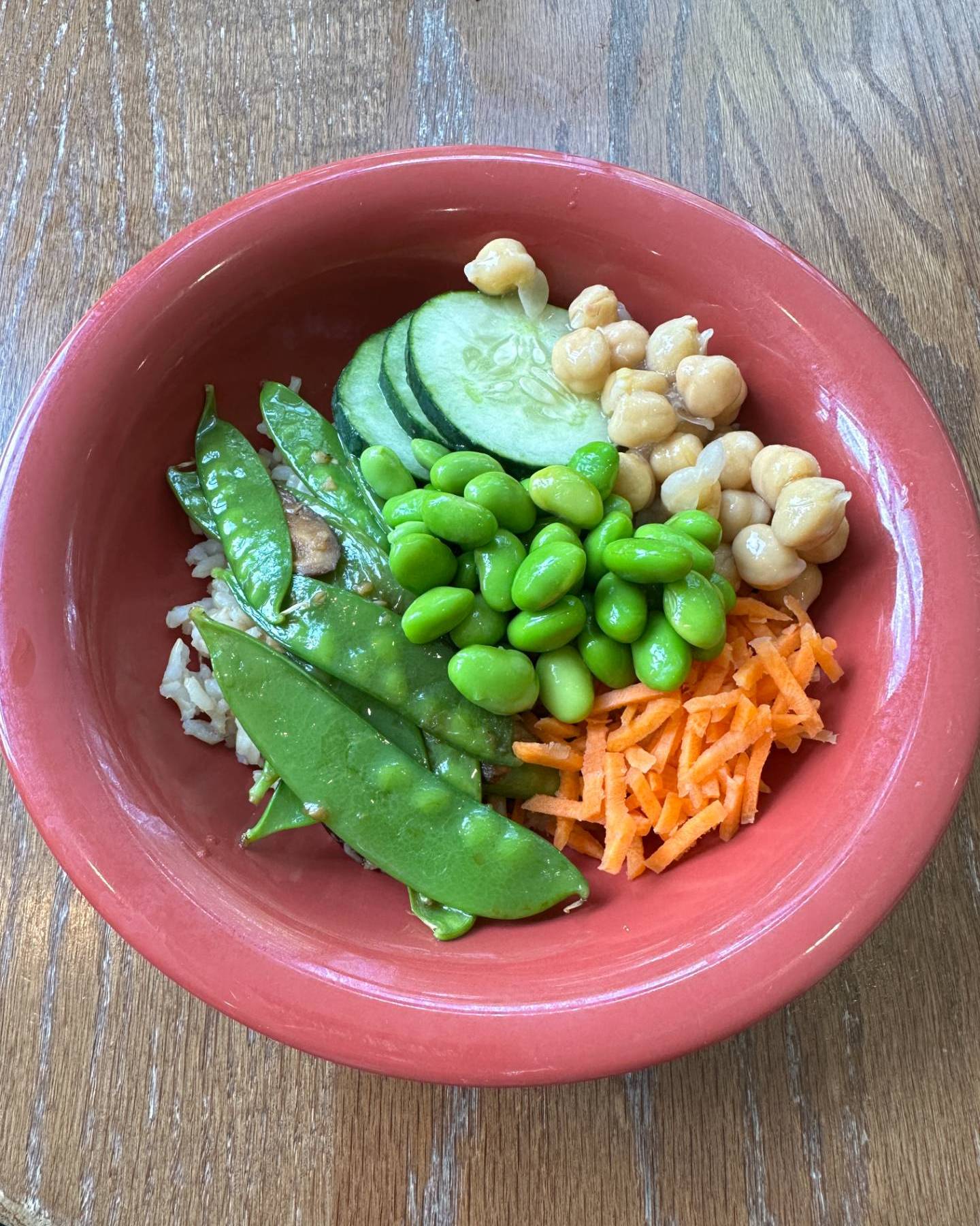 A photograph of a red bowl with chickpeas, cucumbers, edamame, spring peas, shredded carrots, and rice.