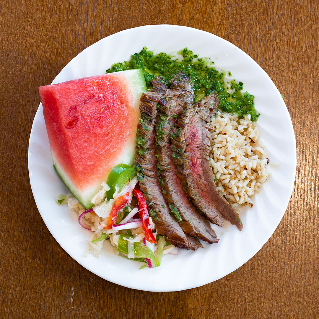 A top down photograph of a white plate with a slice of watermelon, slices of steak, pesto, brown rice, and peppers.