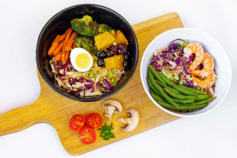 A top-down photograph of two bowls sitting atop a wooden serving board. One bowl contains carrots, brocoli, squash, berries, onions, and rice. The second bowl has quinoa, green beans, shrimp, and red onions.