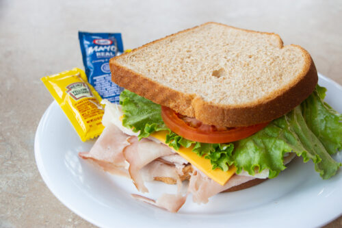 Photograph of a sandwich with turkey, cheese, lettuce, and tomato. A single serve packet of mayo and mustard sit next to it on the plate.