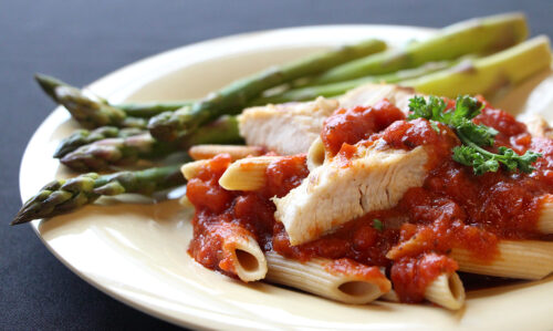 Photograph of wheat pasta with chicken and red sauce and a side of asparagus.