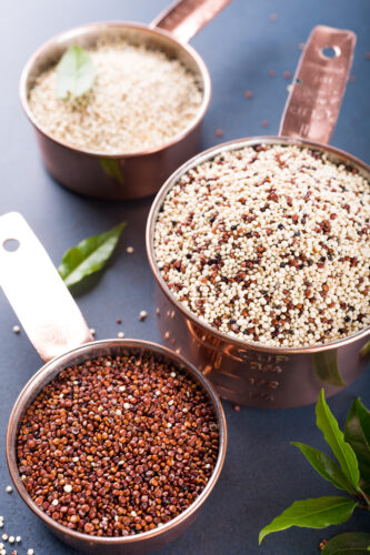 Stack of three Mixed raw quinoa, South American grain, in copper measuring cups with bay laurel leaves on blue background. Healthy and gluten free food.