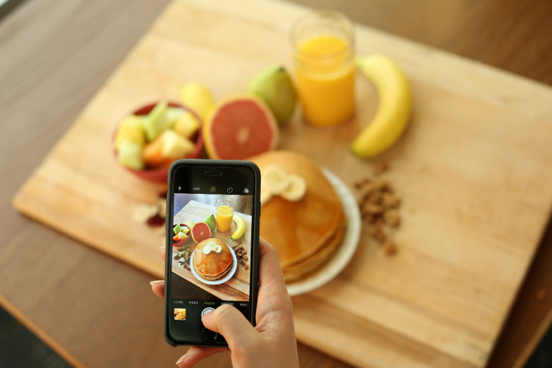 An image of a hand holding an iphone and taking a photo of a tray of food including a plate of pancakes with banana slices, a bowl of fruit, half a grapefruit, a pear, a banana, nuts and raisins, granola, and a cup of orange juice.