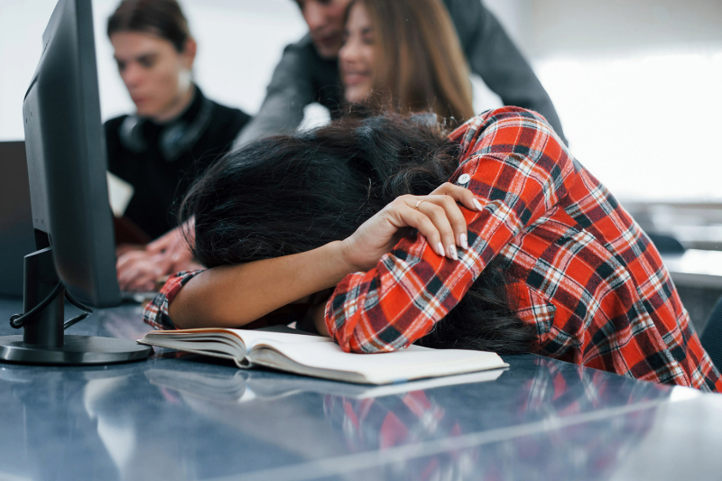 A photograph of a young woman with her head down on a desk.