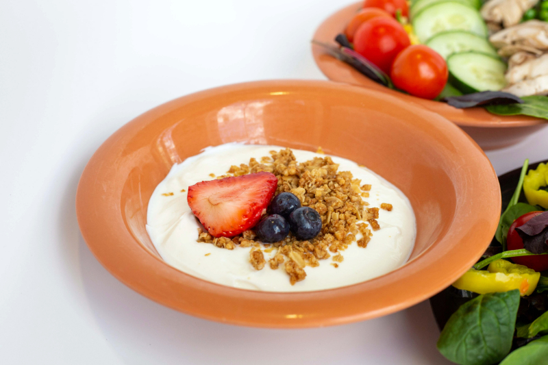 an orange bowl filled with yogurt topped with blueberries, raspberries, and granola. A salad sits off to the side of the image.